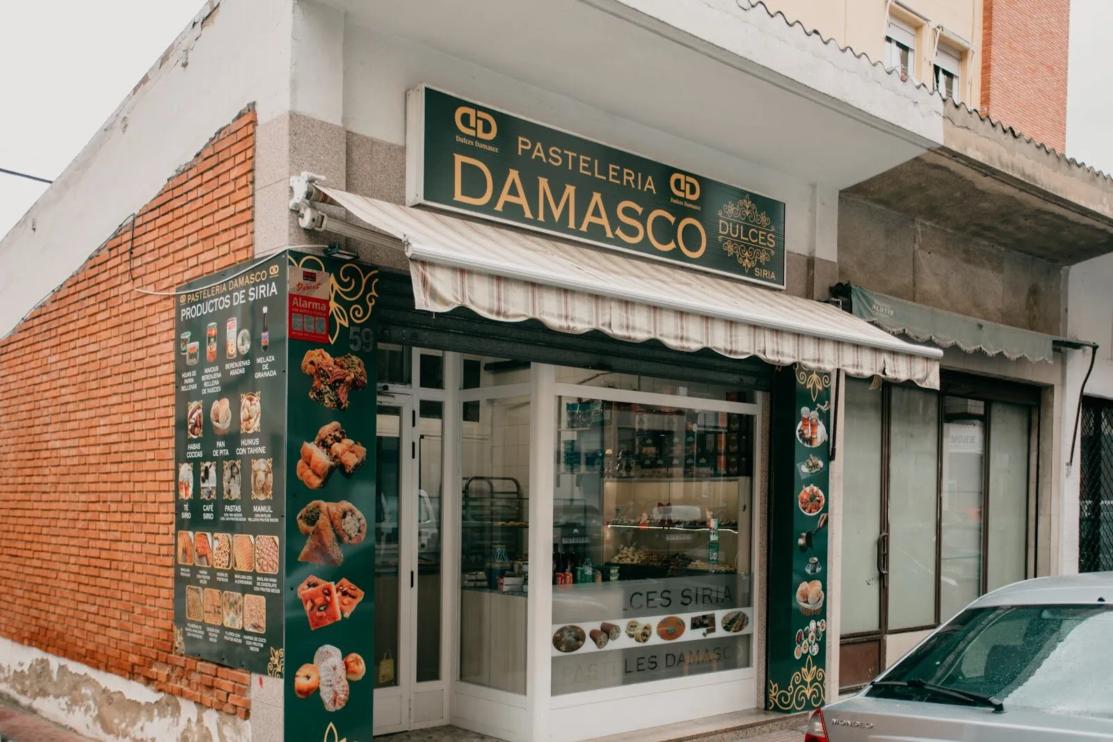 Fachada de Pastelería Damasco: rótulo verde y dorado, toldo de rayas y vinilo con los productos de Siria